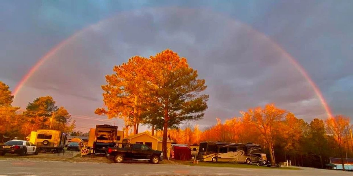 A vibrant rainbow arches over autumn trees and parked vehicles at sunset.
